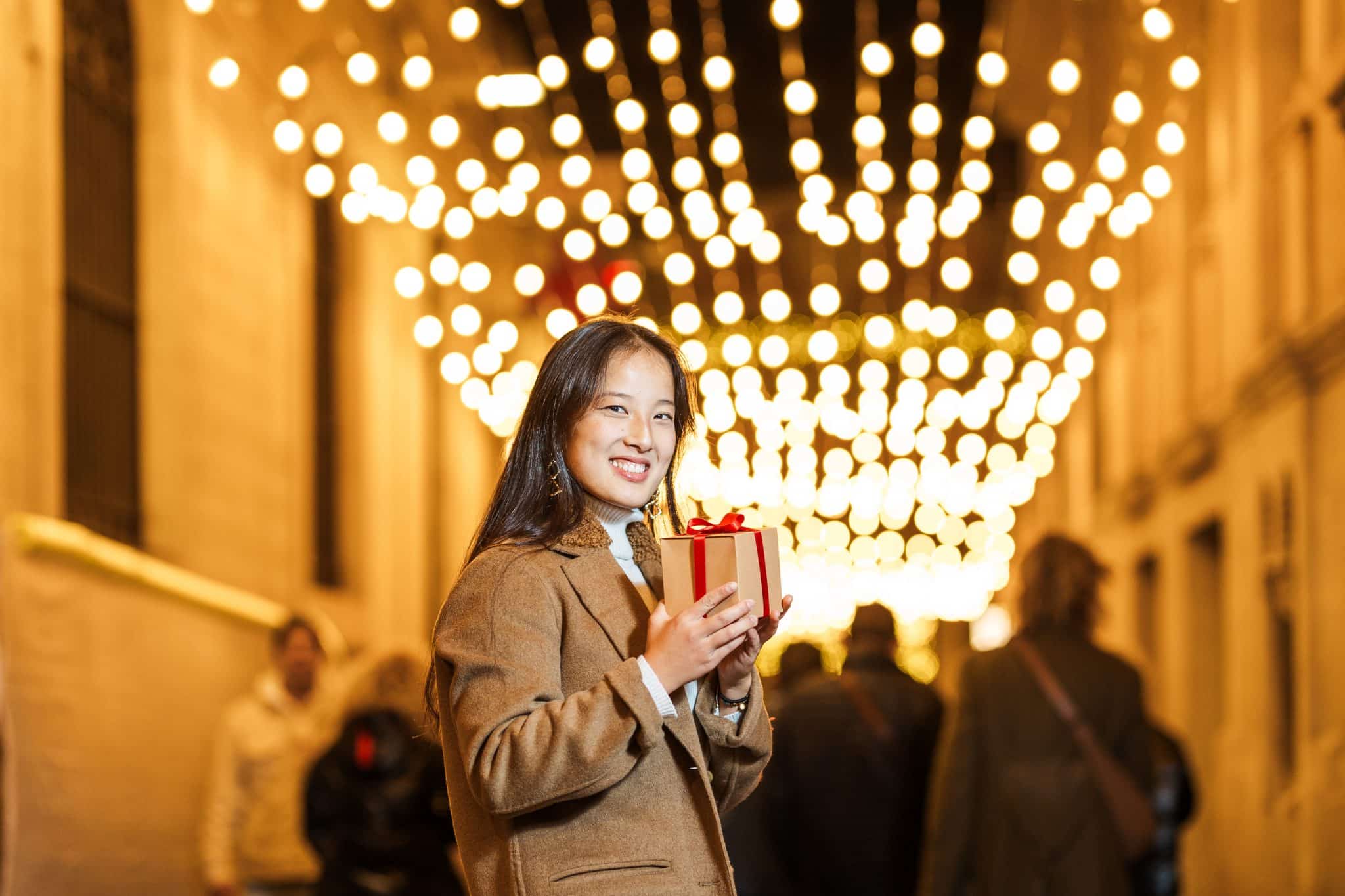 Smiling young woman holding a gift box with red ribbon, standing on a street decorated with festive lights at night